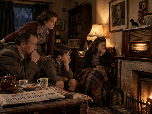 A British family in the early 1940s sitting tensely around the wireless set on the mantelpiece, with a coal fire burning and a newspaper headline reading Britain Stands Firm