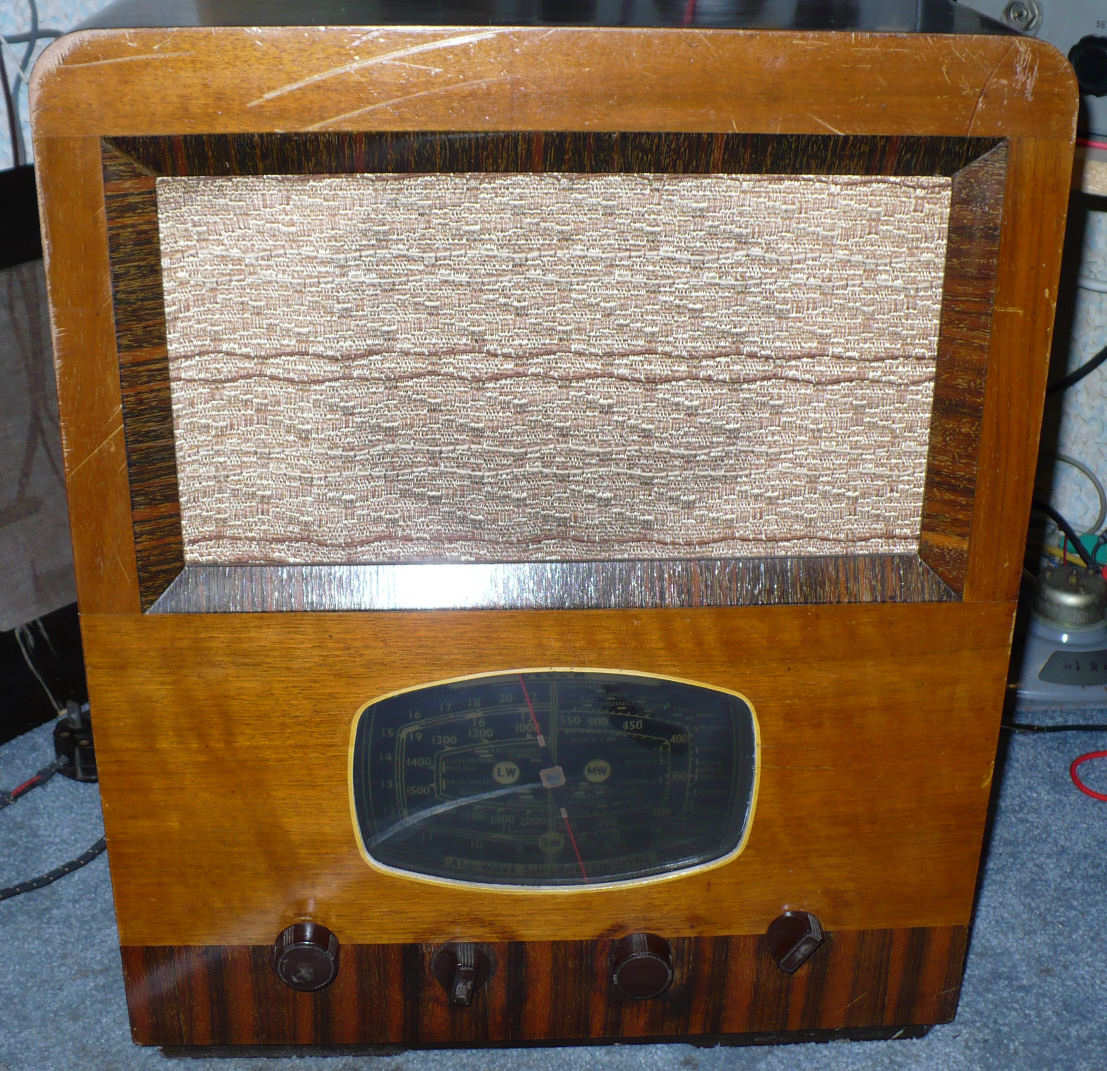 Light walnut upright cabinet with a patterned speaker cloth above a small oval black tuning dial and three knobs