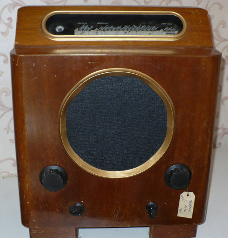 Walnut cabinet on short legs, with a long narrow tuning window at the top, a round speaker with a brass-coloured rim and two black knobs