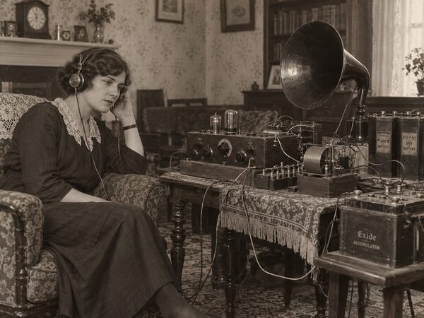 Woman in a 1920s dress sitting in an armchair wearing headphones, beside a table holding a homemade valve receiver, a horn loudspeaker, dry batteries and an Exide accumulator